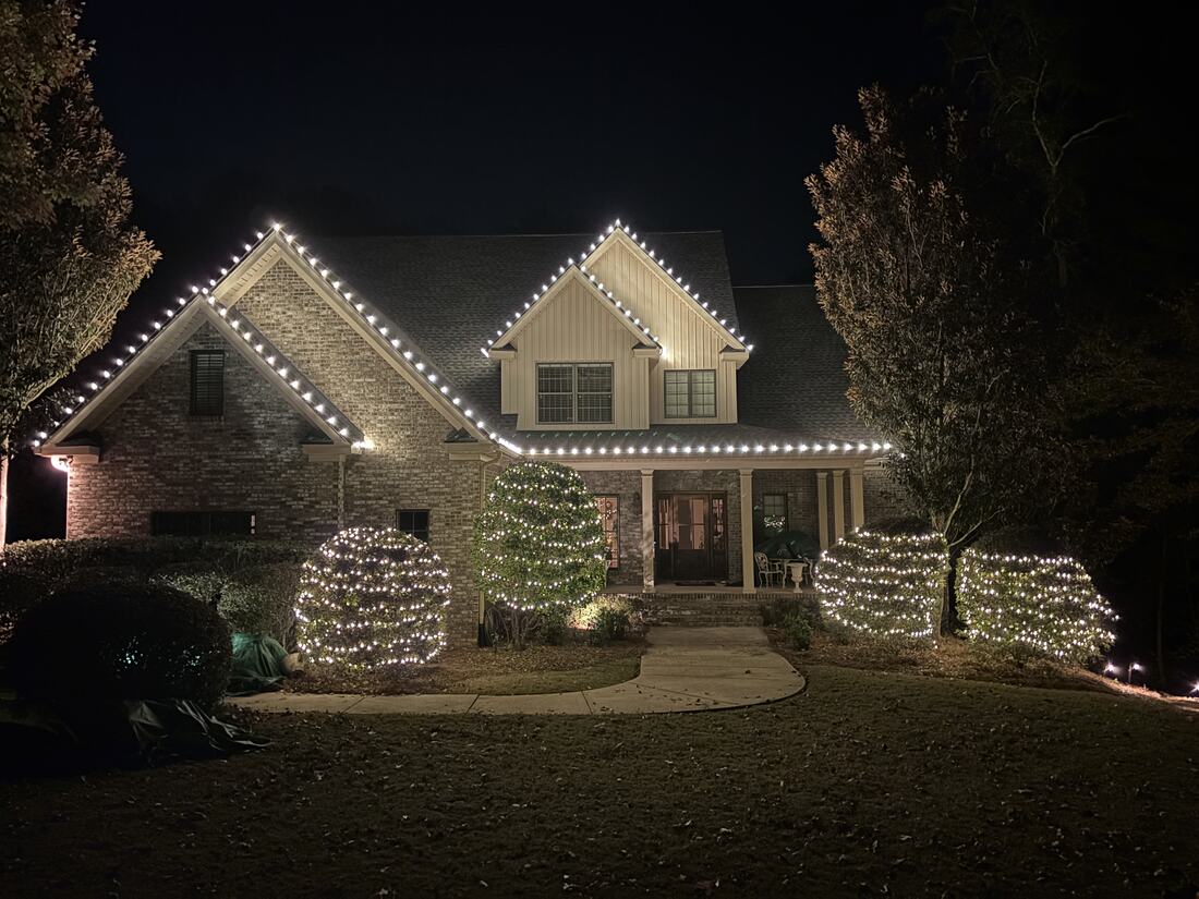 A two-storey brick home at night with roofline lights and net-lit shrubs across the front beds