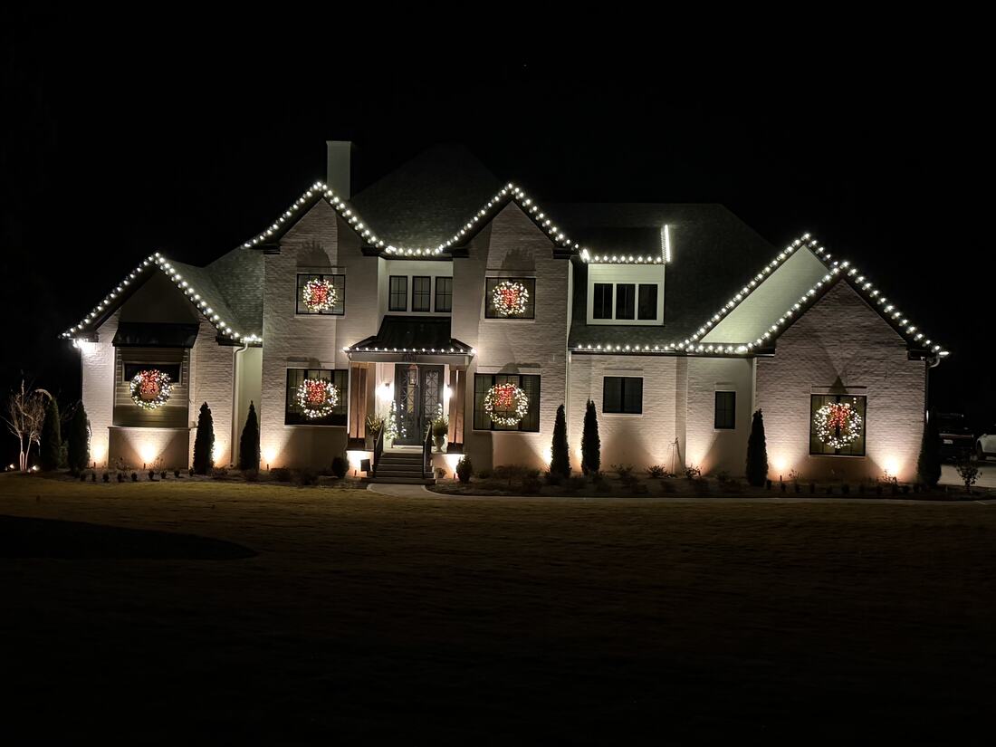 A large brick home at night outlined along every roof angle with warm white lights and lit wreaths