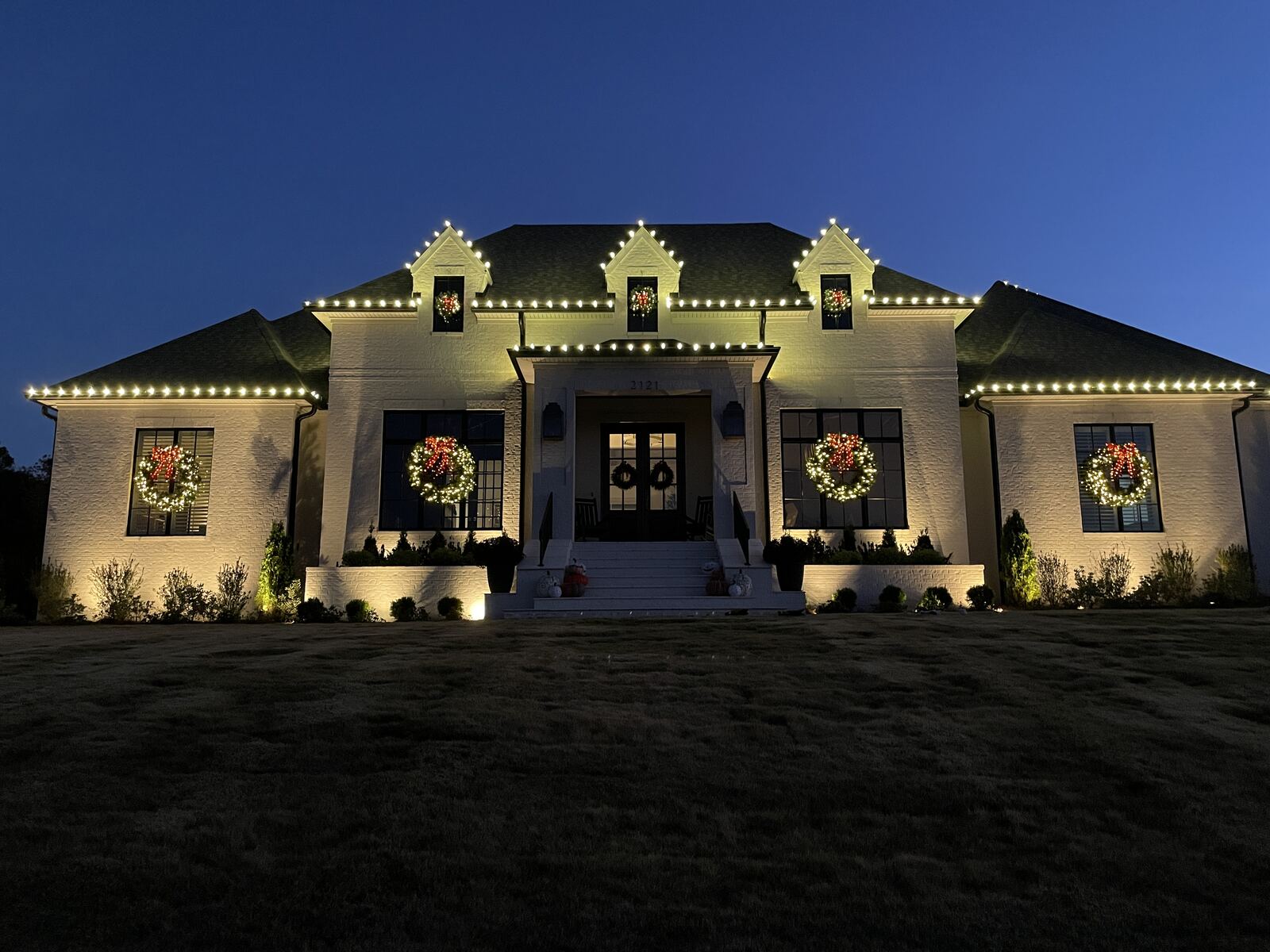 A white brick home at dusk with roofline lights, lit wreaths in five windows, and uplighting across the landscaping